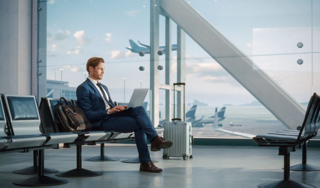 Modernes Flughafen-Terminal: Gutaussehender Geschäftsmann arbeitet am Laptop, während er auf seinen Flug wartet. Mann sitzt in einer Boarding Lounge einer großen Fluggesellschaft Hub mit Flugzeugen abfliegen und ankommen