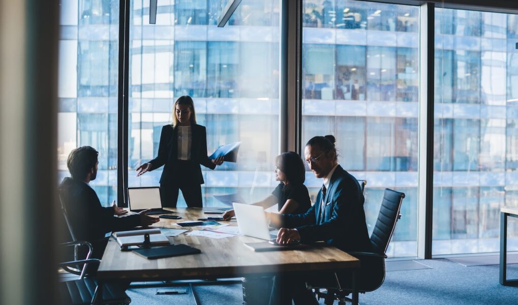 Businessmeeting mit vier Personen in einem modernen Büro mit Glasfassade, eine Frau steht und präsentiert