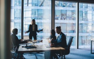 Businessmeeting mit vier Personen in einem modernen Büro mit Glasfassade, eine Frau steht und präsentiert