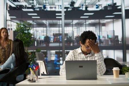 Gestresster Mann sitzt im modernen Buero vor seinem Laptop und haelt sich den Kopf